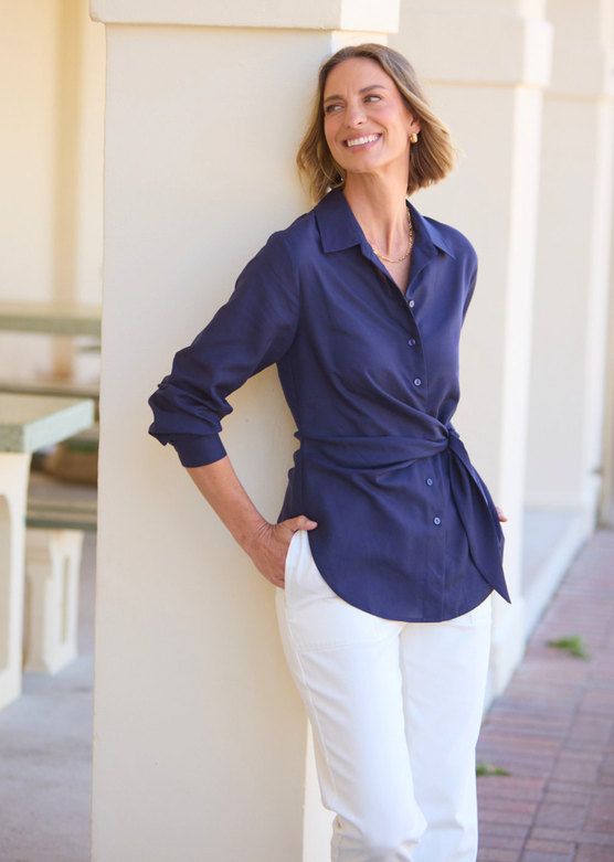 A woman wearing a navy blue long-sleeve button-up shirt tied at the waist and white pants, leaning against a cream-colored architectural column in an outdoor setting.